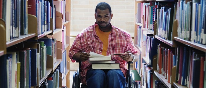 A student in a wheelchair reads books in a library aisle. 